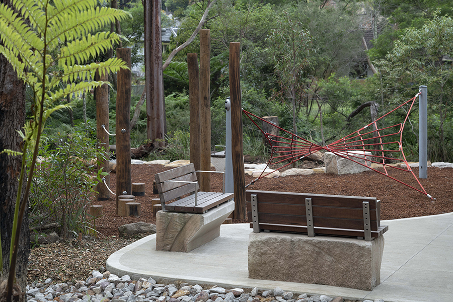 A play area made from timber in Nandi Reserve. The floor is wood chips and there is a rope climbing frame, and several benches for seating.