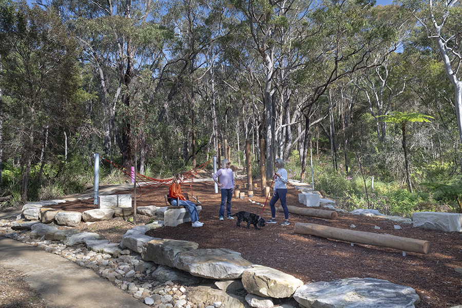 Three people and a dog standing in the new play area in Nandi Reserve. The play area has timber play equipment and one rope climbing frame. The play surface is woodchips.