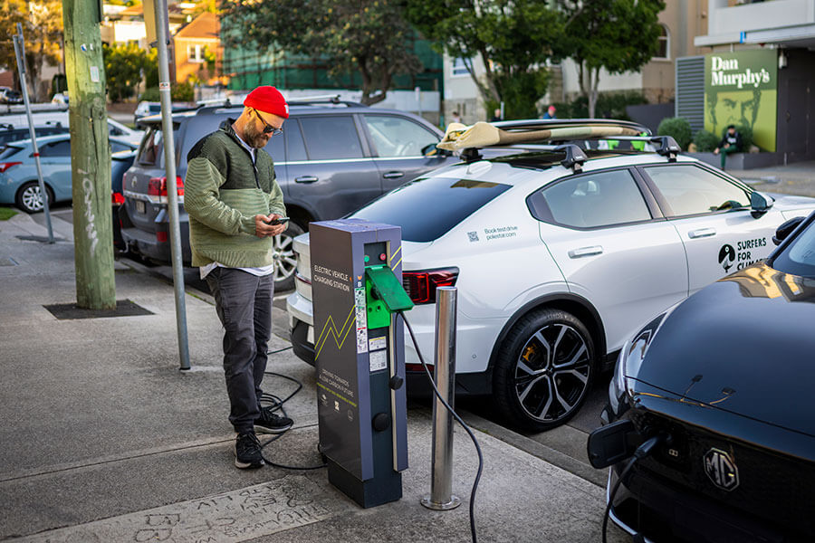 A person wearing black jeans, a green jumper, a red beanie and sunglasses, standing on a footpath next to an electric vehicle charging station. Credit: Jessica Lindsay / Department of Climate Change, Energy, the Environment and Water