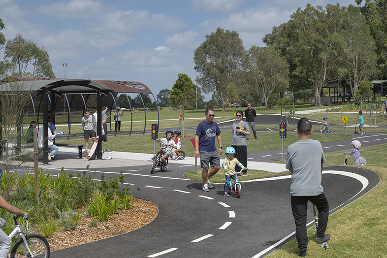 Children riding bikes on a bike path in a large park. There are families in the background sitting under shaded shelter, trees and children playing.