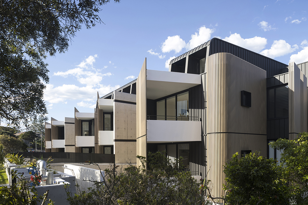 A modern, mid-rise apartment block on a leafy street. The building has curved, slatted corners, corrugated iron enclosed balconies and street-facing balconies with timber privacy screens.